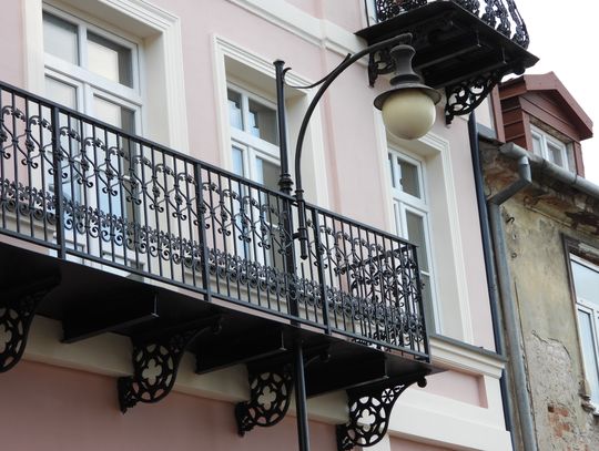 Przez środek balkonu w centrum miasta przechodzi latarnia. Mieszkaniec pyta kto pozwolił ją postawić na środku chodnika 